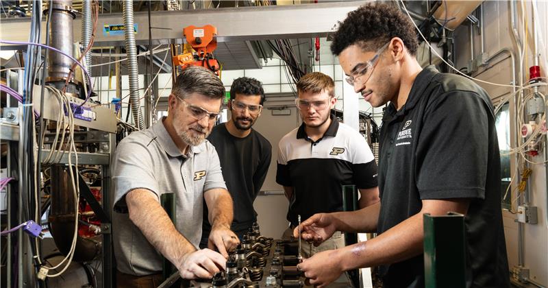 Four Purdue researchers with safety goggles stand in a crowded workspace, two of them working on a large engine.