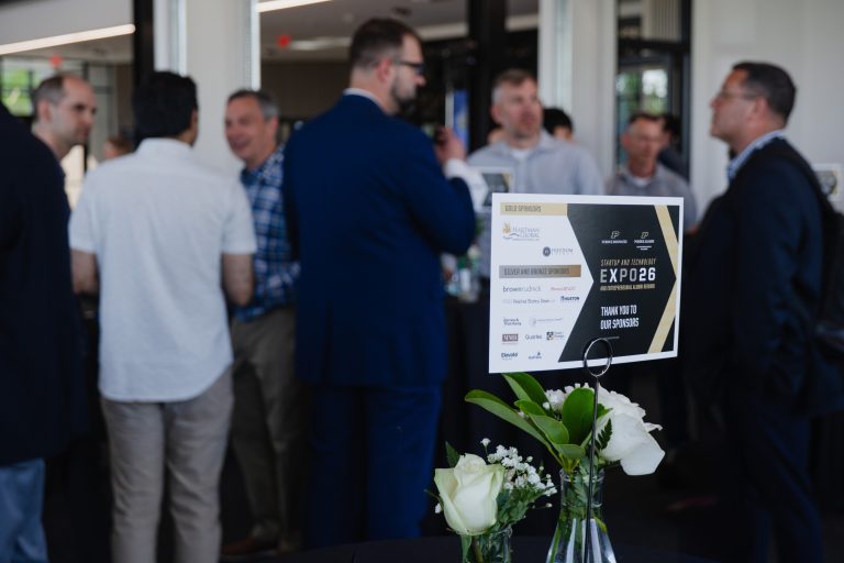 Group of professionals chatting at a corporate event; sponsor sign with logos and white roses on table in foreground.