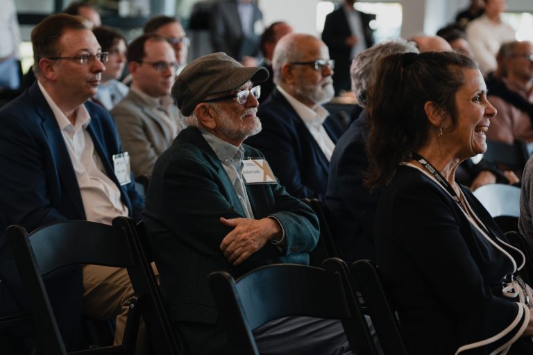 Audience at a conference, man with gray cap and glasses in foreground among seated attendees with badges