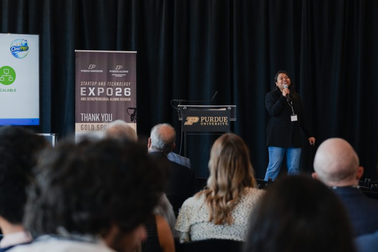 Female presenter on stage speaking into a handheld microphone with a Purdue Expo 26 banner in the background, audience in front. This is at Purdue University.
