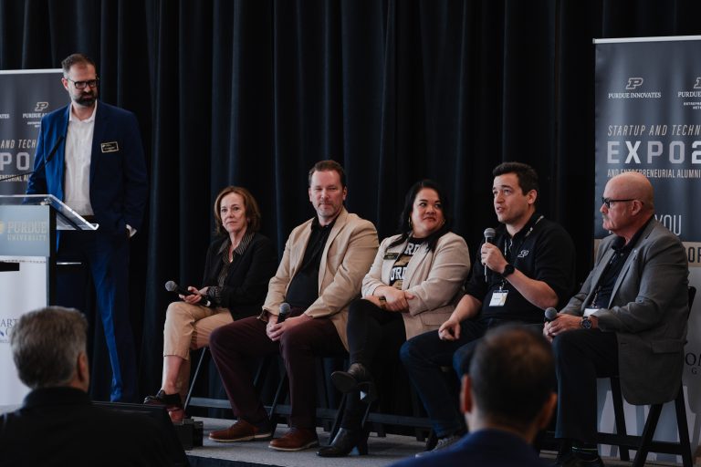 Panelists seated on stage during a Purdue startup expo, with a speaker at a podium on the left.