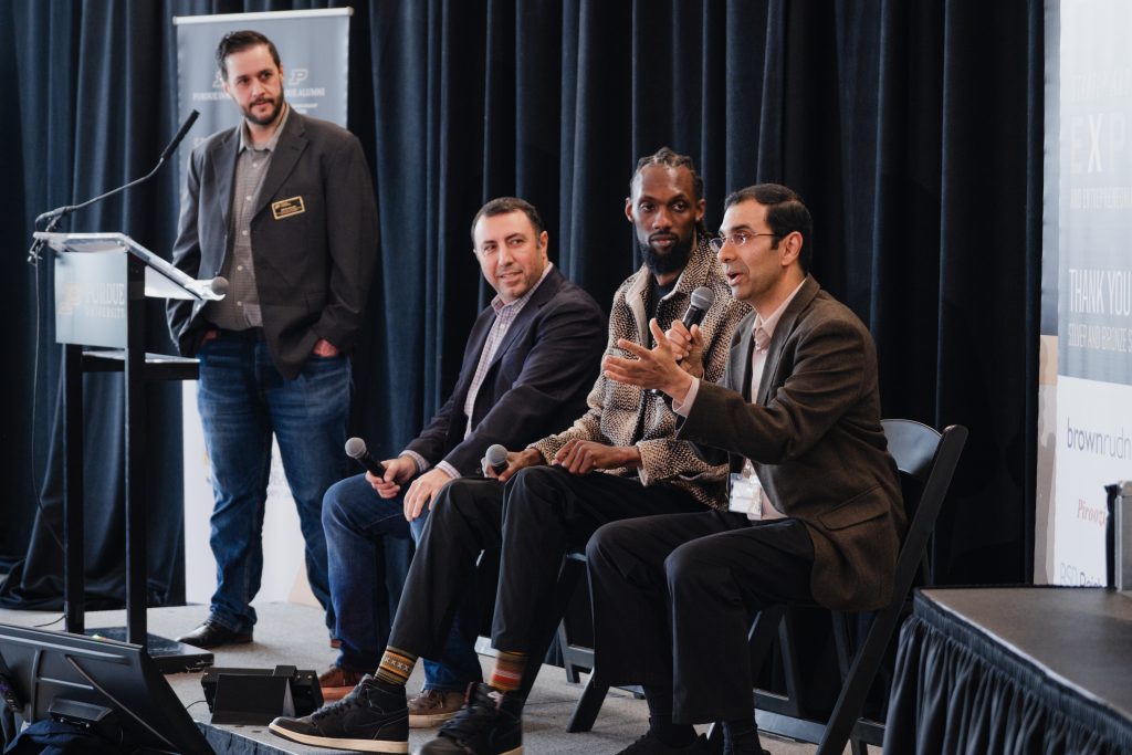 Panel discussion with four men on a stage, one speaking into a handheld mic as others listen, conference banners in the background.