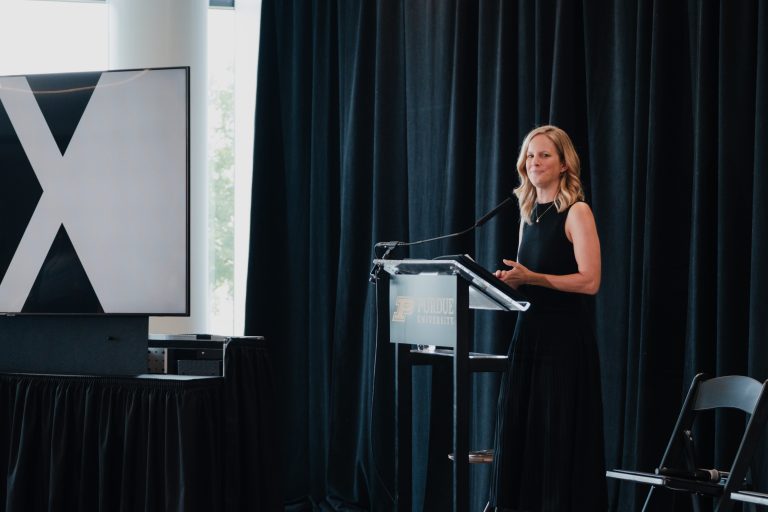 Woman in a black dress giving a presentation at a podium with Purdue University branding, dark curtains behind her.