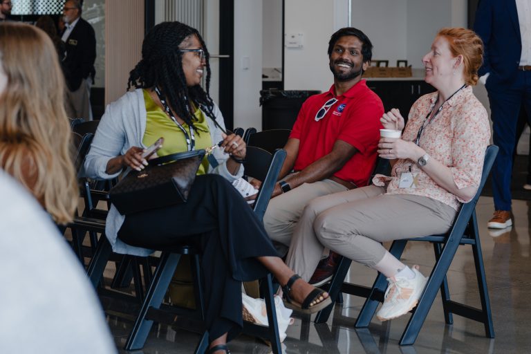 Three people seated in folding chairs, smiling and talking in a bright, informal meeting space.