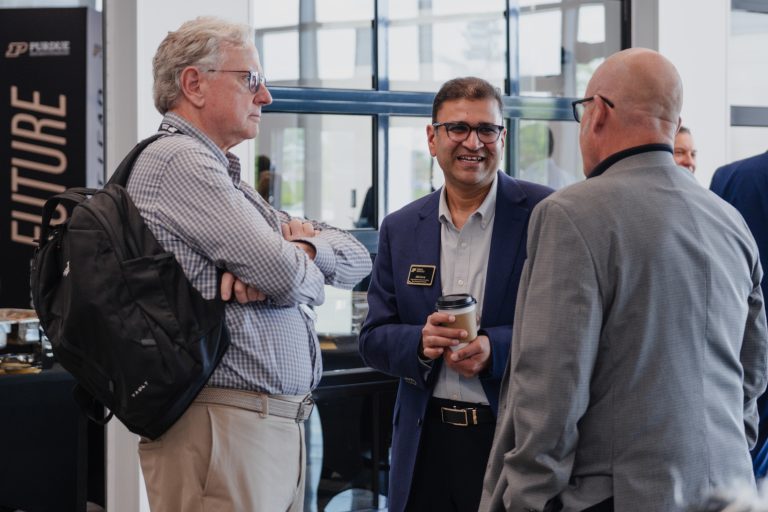 Three men in business attire chat in a bright lobby; one with a backpack, another in a blue blazer holding a coffee cup, and a third facing away in a gray suit.