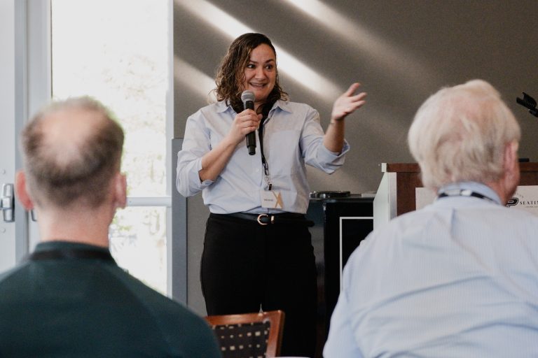 Woman in a light blue blouse speaks into a handheld microphone to an audience, gesturing with her free hand in a conference room.