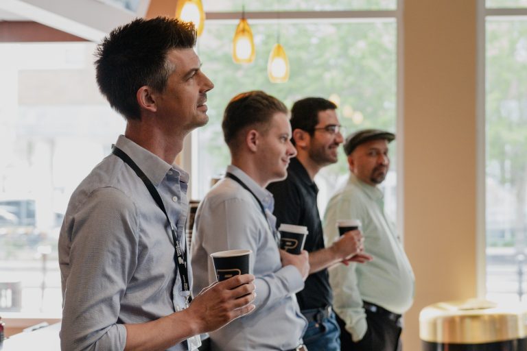 Group of professionals in a bright lobby, standing in a line with takeaway coffee cups.