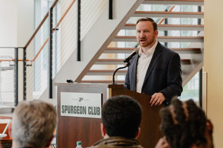 Man in a suit delivering a speech at a podium in a club setting, with a 'SPURGEON CLUB' sign visible on the lectern