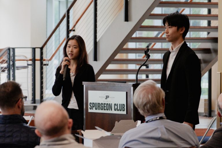Two presenters stand at a podium labeled 'Spurgeon Club' addressing an audience in a modern space with a staircase behind them