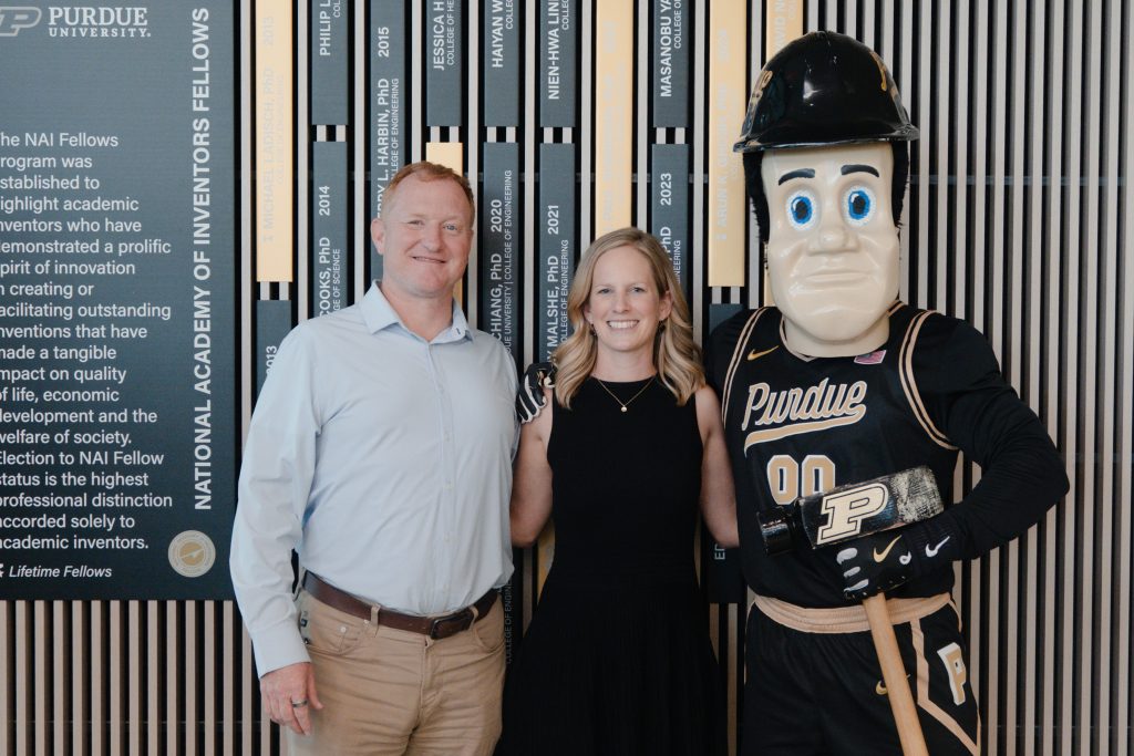 Man in light blue shirt and khaki pants poses with a woman in black, next to the Purdue Boilermakers mascot in a striped backdrop.