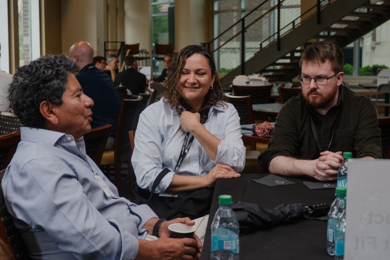 Three colleagues sit at a table in a bright indoor space, with the woman in the center smiling as the two men talk nearby.] ,