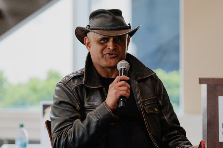Man in a leather jacket and black cowboy hat speaks into a handheld microphone, likely at an event.