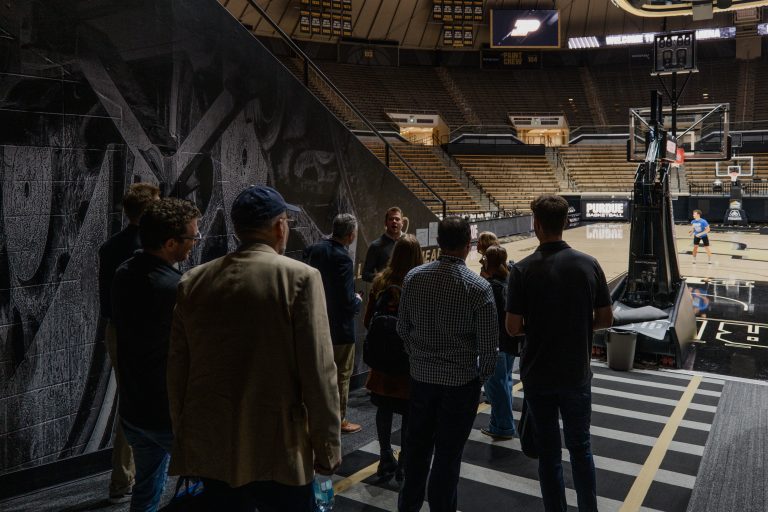 Group of people stand in a tunnel or backstage area overlooking a basketball court in a large arena, with a mural-covered wall on the left and empty bleachers in the background.