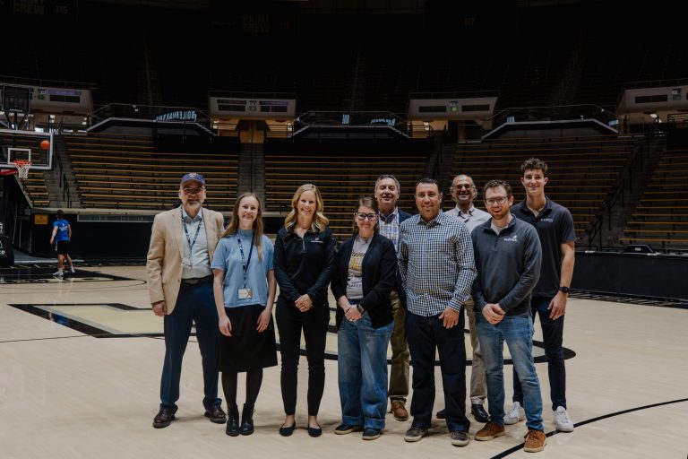 Nine adults stand together on a basketball court inside an arena, smiling for a group photo with empty bleachers in the background.