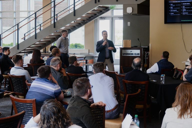 Speaker holding microphone addresses group in a conference room, presenting near a lectern labeled 'Spurgeon Club'