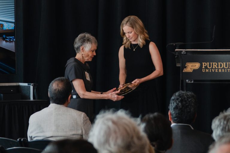 Older woman presents a plaque to a younger woman on a stage at Purdue University award ceremony