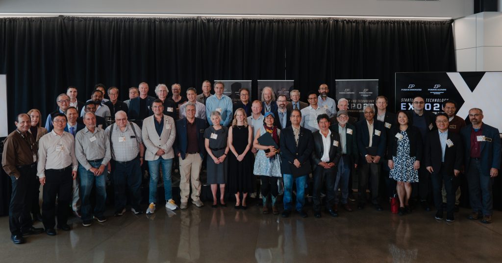 Group of diverse professionals posing for a group photo at a conference, standing on a stage with black curtains and banners behind them, name tags visible.