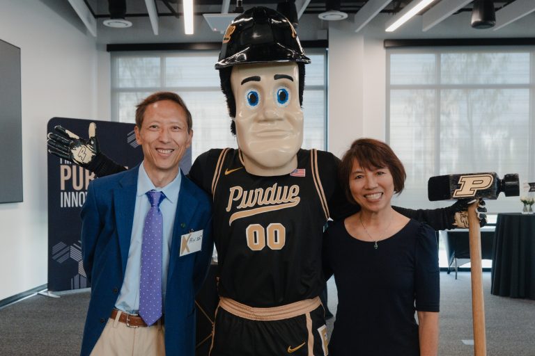 Smiling man in a blue suit and purple tie stands beside Purdue's mascot in a modern office setting with a woman partially visible on the right.