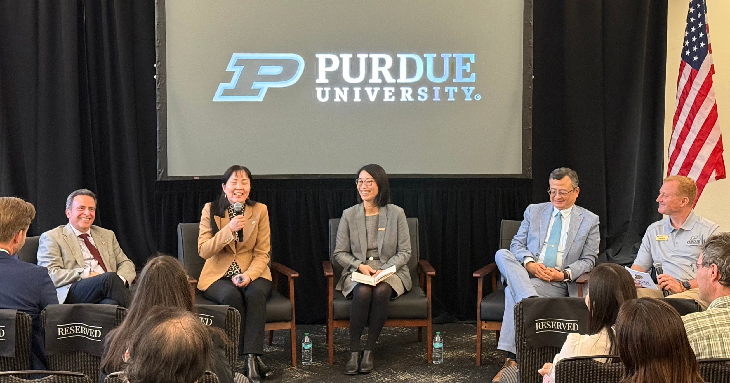 A smiling group of five sits in a semicircle for a fireside chat. Behind them is a Purdue University logo on a large screen.
