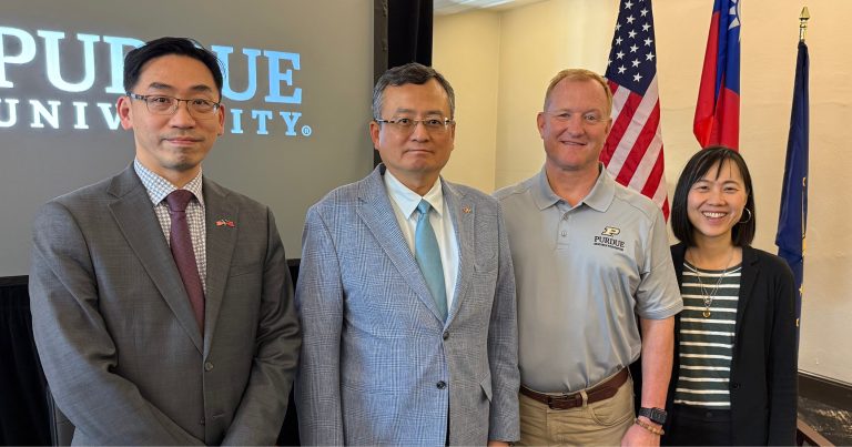 Four professionals pose for a photo at a Purdue University event, with the Purdue sign and flags in the background.