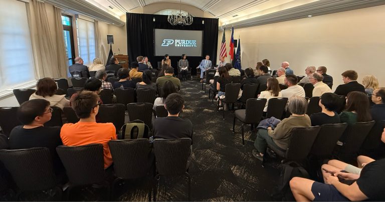 Panel discussion in a Purdue University event room with speakers on stage, audience seated facing the panel, flags to the side, and a screen displaying the Purdue logo.