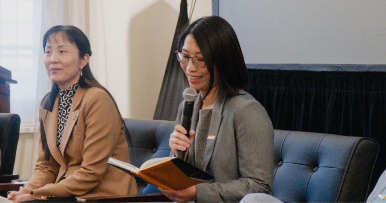 Two women seated on a panel; one speaks into a microphone while holding an open book on her lap.