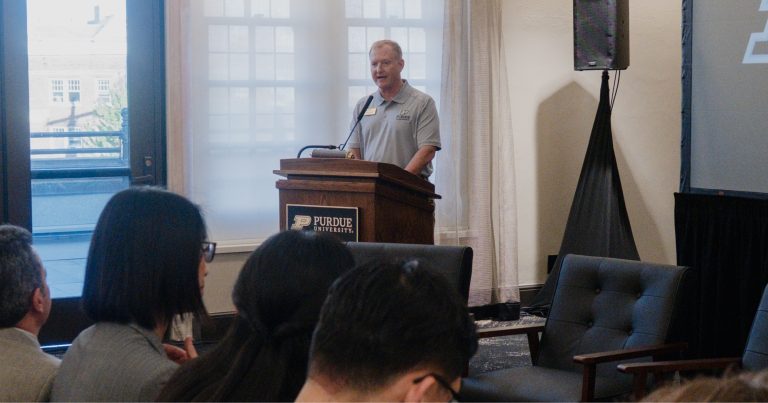 Man at a wooden podium giving a talk at a Purdue University event, with an audience in the foreground.