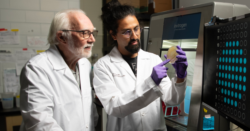 Two researchers in white lab coats examine a petri dish beside a large Invitrogen biomedical instrument in a lab.
