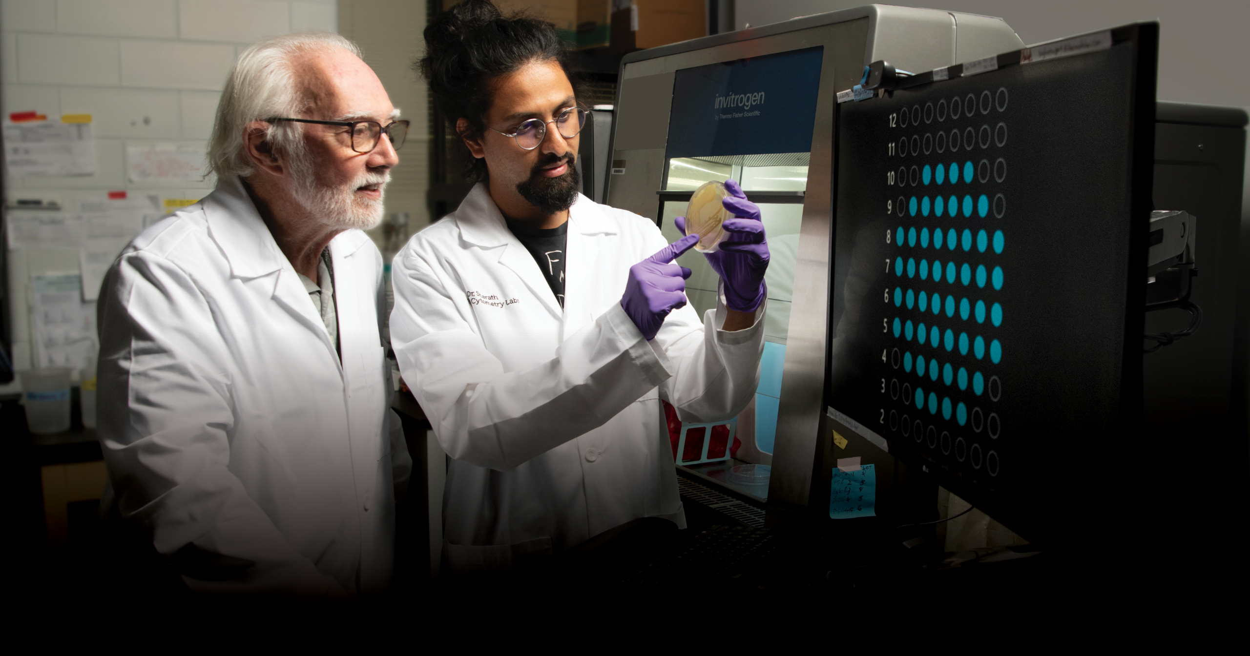Two researchers in white lab coats examine a petri dish near a scientific machine in a lab.