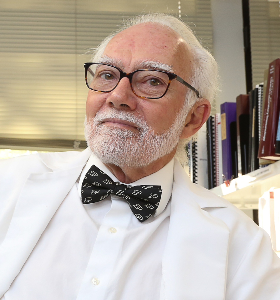 Portrait of an elderly man with white hair and beard wearing glasses, a white lab coat, and a patterned bow tie in an office setting