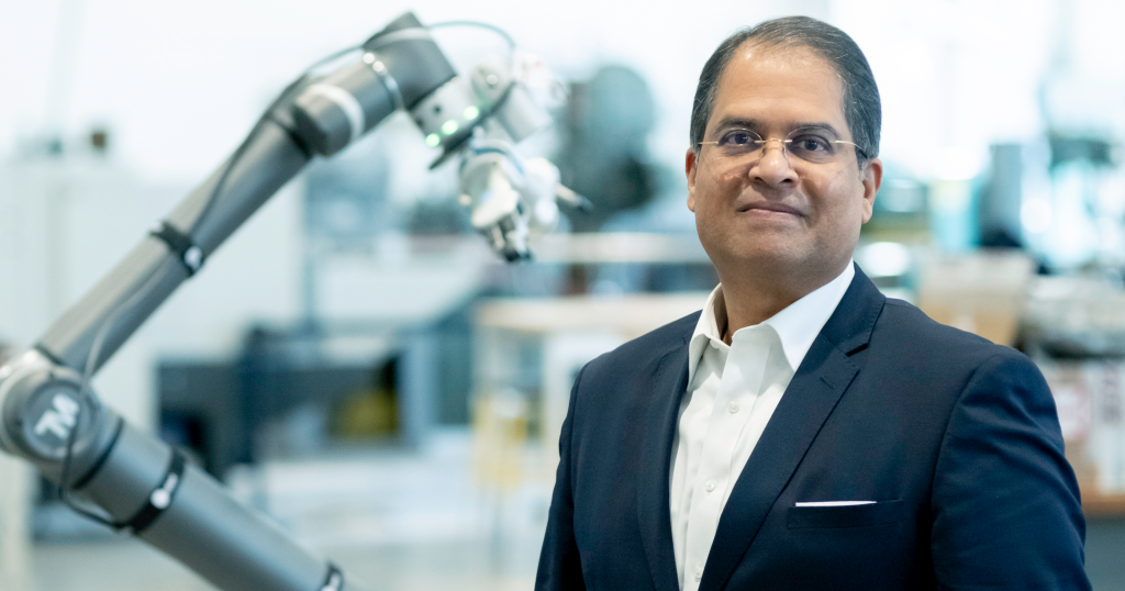A headshot of Ajay Malshe wearing a suit. He stands in a lab setting, a robotic arm slightly out of focus behind him.