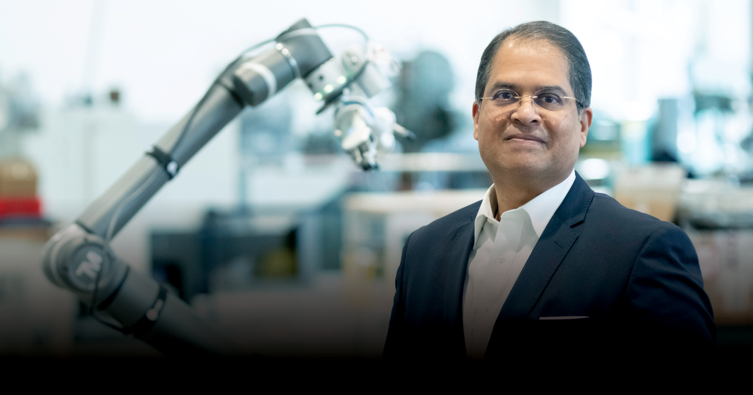 A headshot of Ajay Malshe wearing a suit. He stands in a lab setting, a robotic arm slightly out of focus behind him.