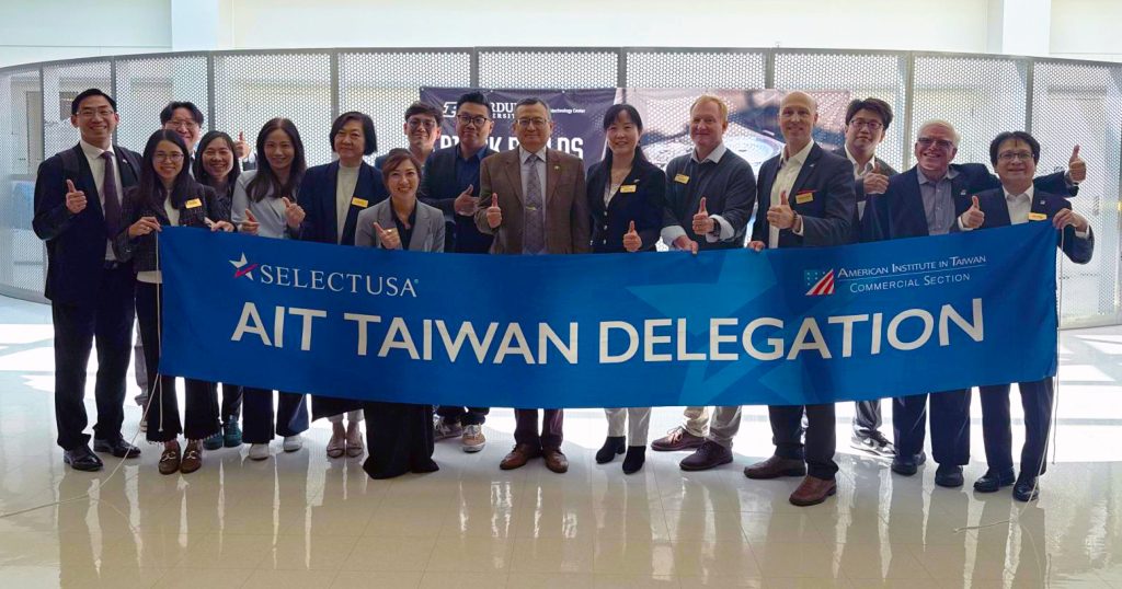 A group stands in a large white hall, giving a thumbs up with a large sign that reads SelectUSA AIT Taiwan Delegation