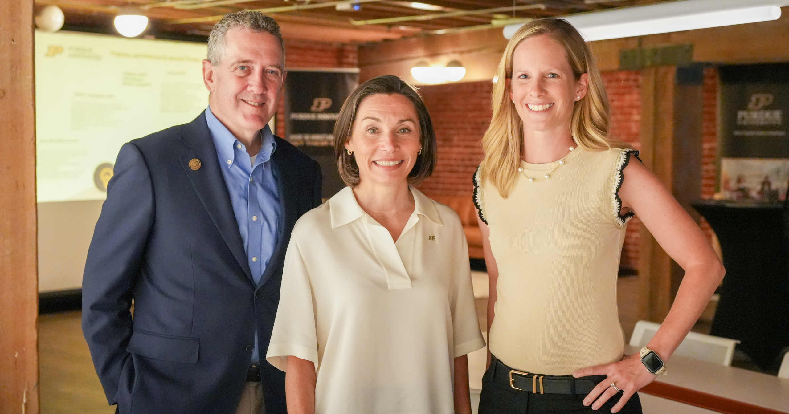 Dean Jim Bullard, Dean Lucy Flesch, and Purdue Innovates SVP Brooke Beier stand close together, smiling, in an open brick workspace.