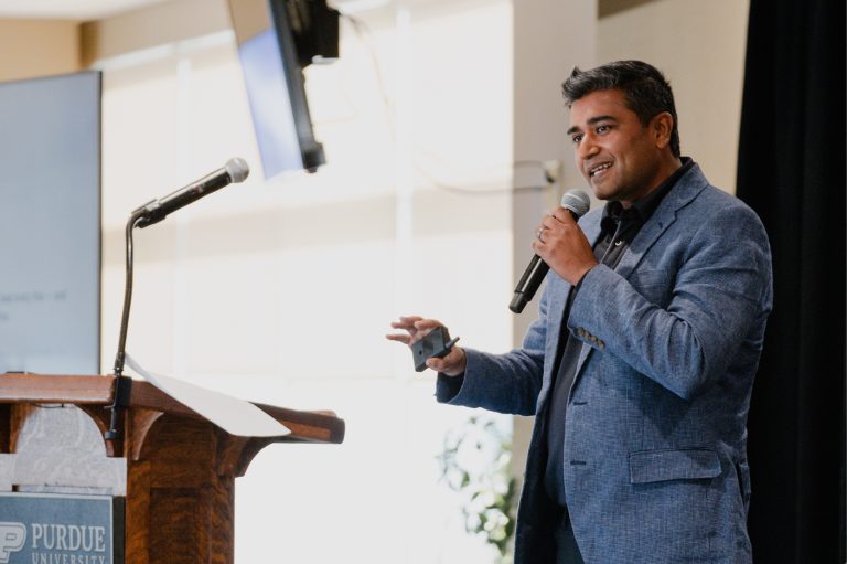 Speaker addresses an attentive audience with a handheld microphone beside a wooden podium, against black curtains in a formal presentation setting.