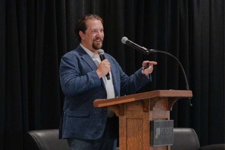 Speaker addresses an attentive audience with a handheld microphone beside a wooden podium, against black curtains in a formal presentation setting.