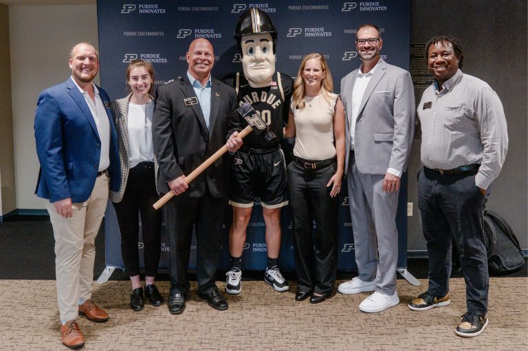 The Purdue Innovates team pose smiling with Purdue Pete, Purdue University’s mascot, at an event; the group stands before a branded backdrop.