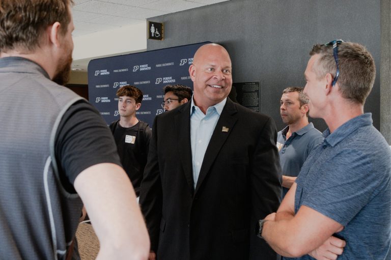 Smiling man in a black suit speaks with two men at an event, with other attendees in the background.