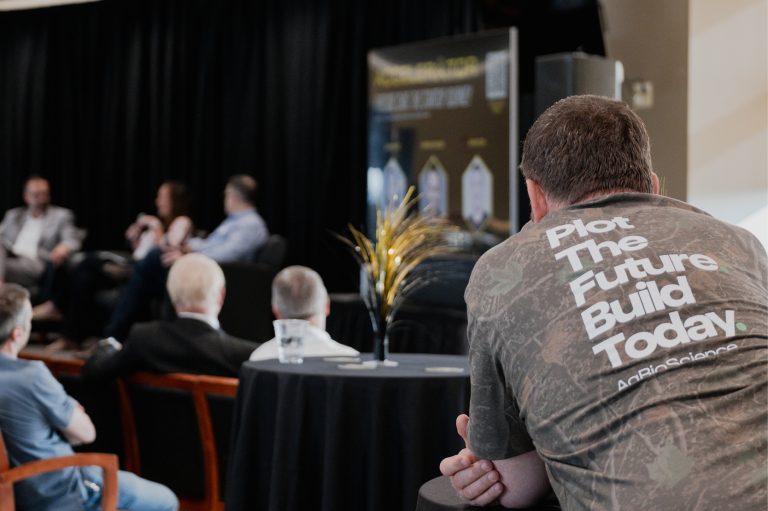 A man with a "Plot the future. Build today." t-shirt, the only thing in focus, stands and watches a presentation on stage.