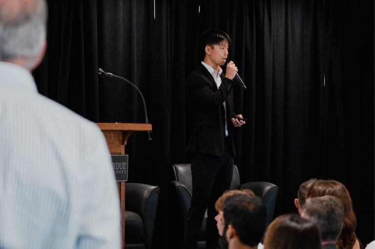 Speaker addresses an attentive audience with a handheld microphone beside a wooden podium, against black curtains in a formal presentation setting.