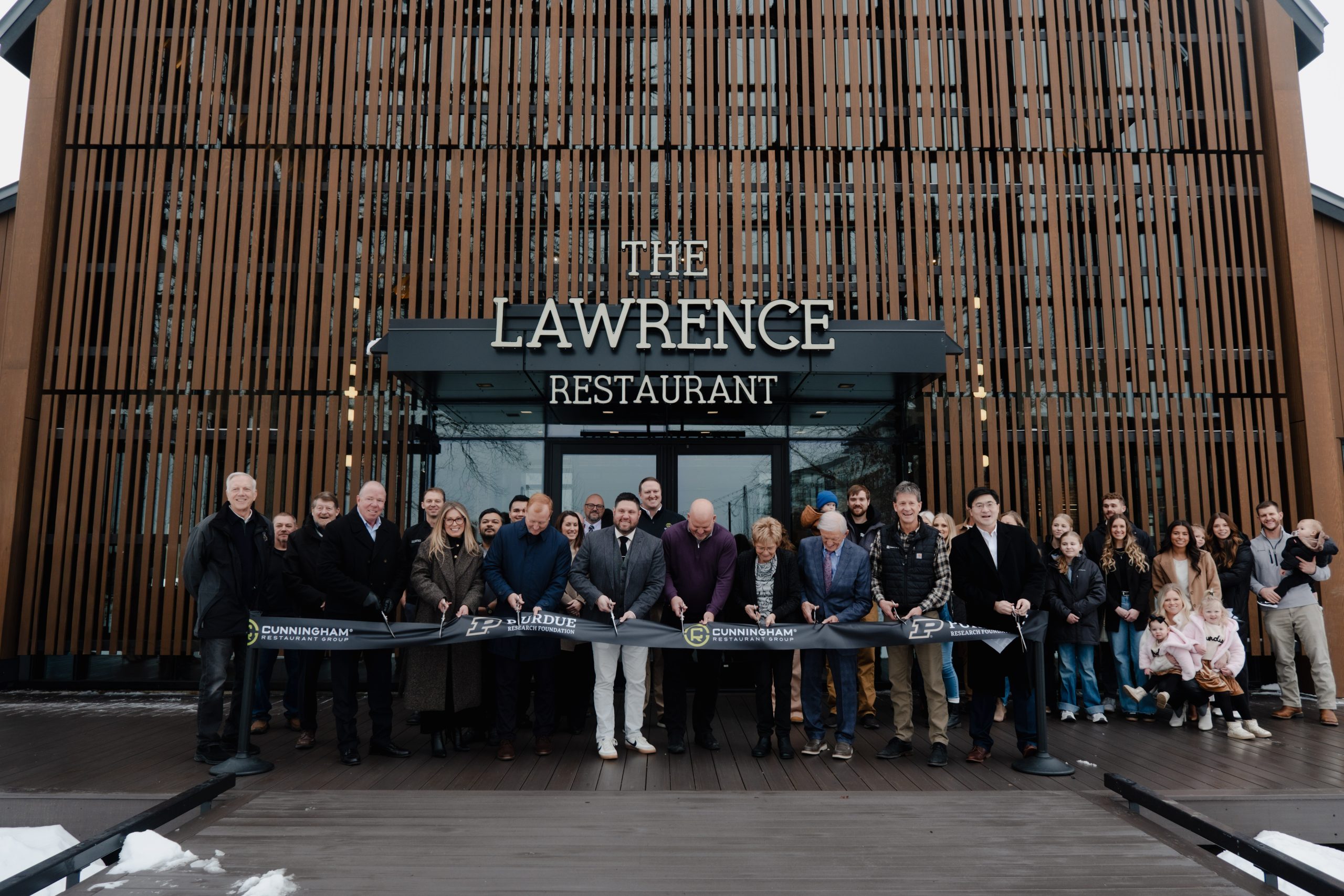 A large group of people stand, mid-ribbon-cutting, in front of the large wood and glass facade of The Lawrence Restaurant