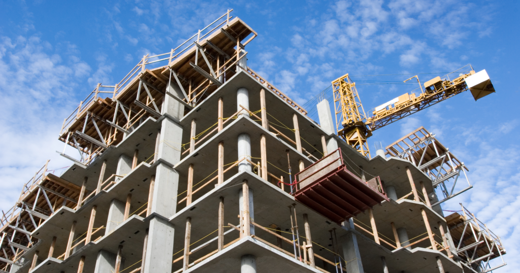 Concrete multi‑story building under construction with exposed floors, wooden safety railings, and scaffolding. A yellow tower crane extends above the structure against a blue, partly cloudy sky.