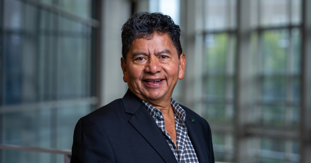 Portrait of a smiling man wearing a dark blazer and checkered shirt, standing in a glass-walled office corridor.