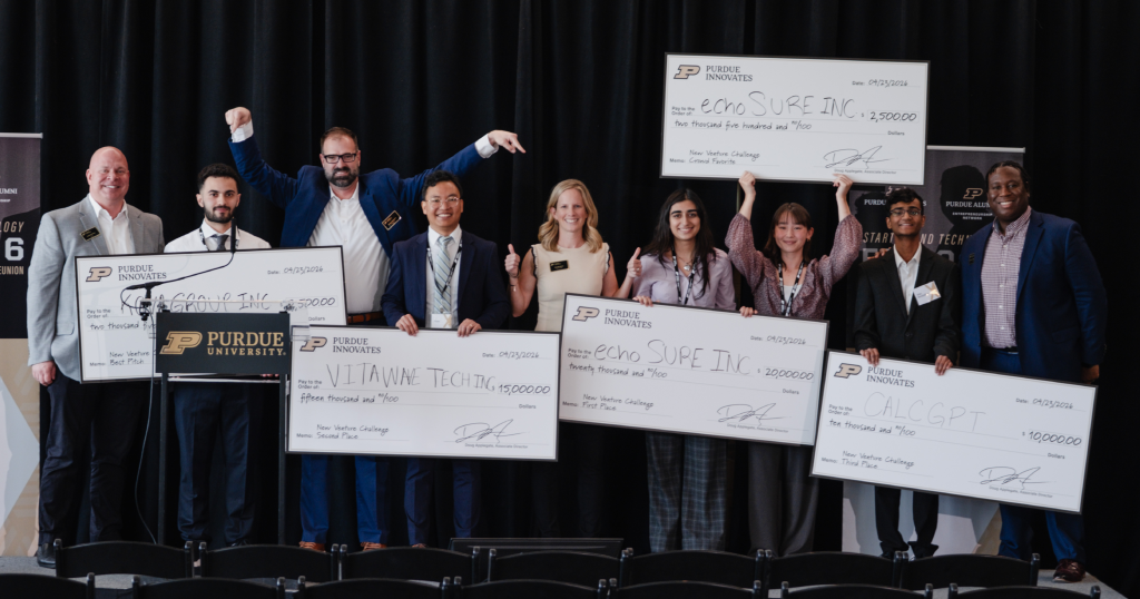 Group of diverse finalists on stage holding oversized checks and smiling at a Purdue Innovates event.
