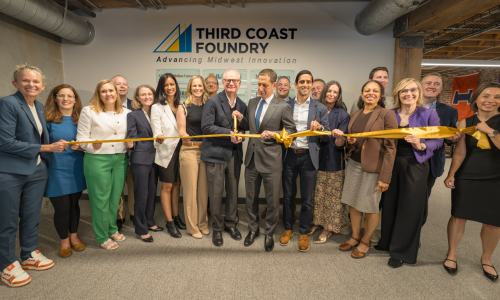A large group of smiling people stand in a semicircle in front of a Third Coast Foundry logo. Two men hold large scissors and are cutting a gold ribbon.