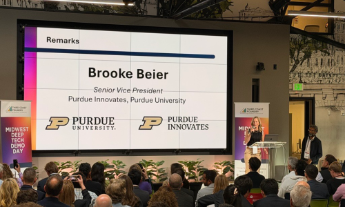 A woman speaks at a podium in front of a large screen displaying "Brooke Beier, Senior Vice President, Purdue Innovates, Purdue University," to an attentive audience.