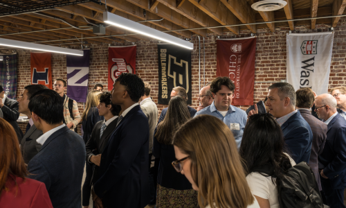 A diverse group of people in business attire engage in lively conversation at a networking event. University banners adorn the brick wall, under exposed beams.