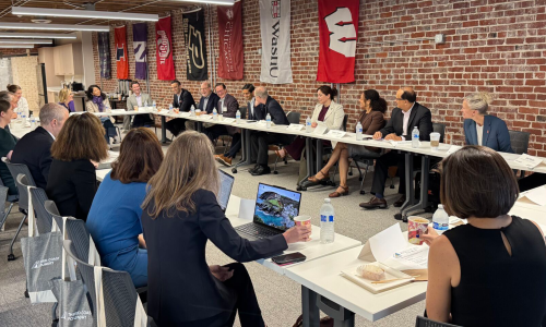 A group of professionals seated around a conference table, engaged in discussion. University banners hang on a brick wall.