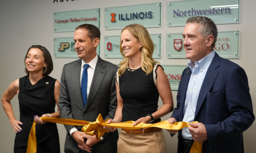 Four people stand side by side holding a yellow ribbon, smiling. Behind them are signs of various universities.